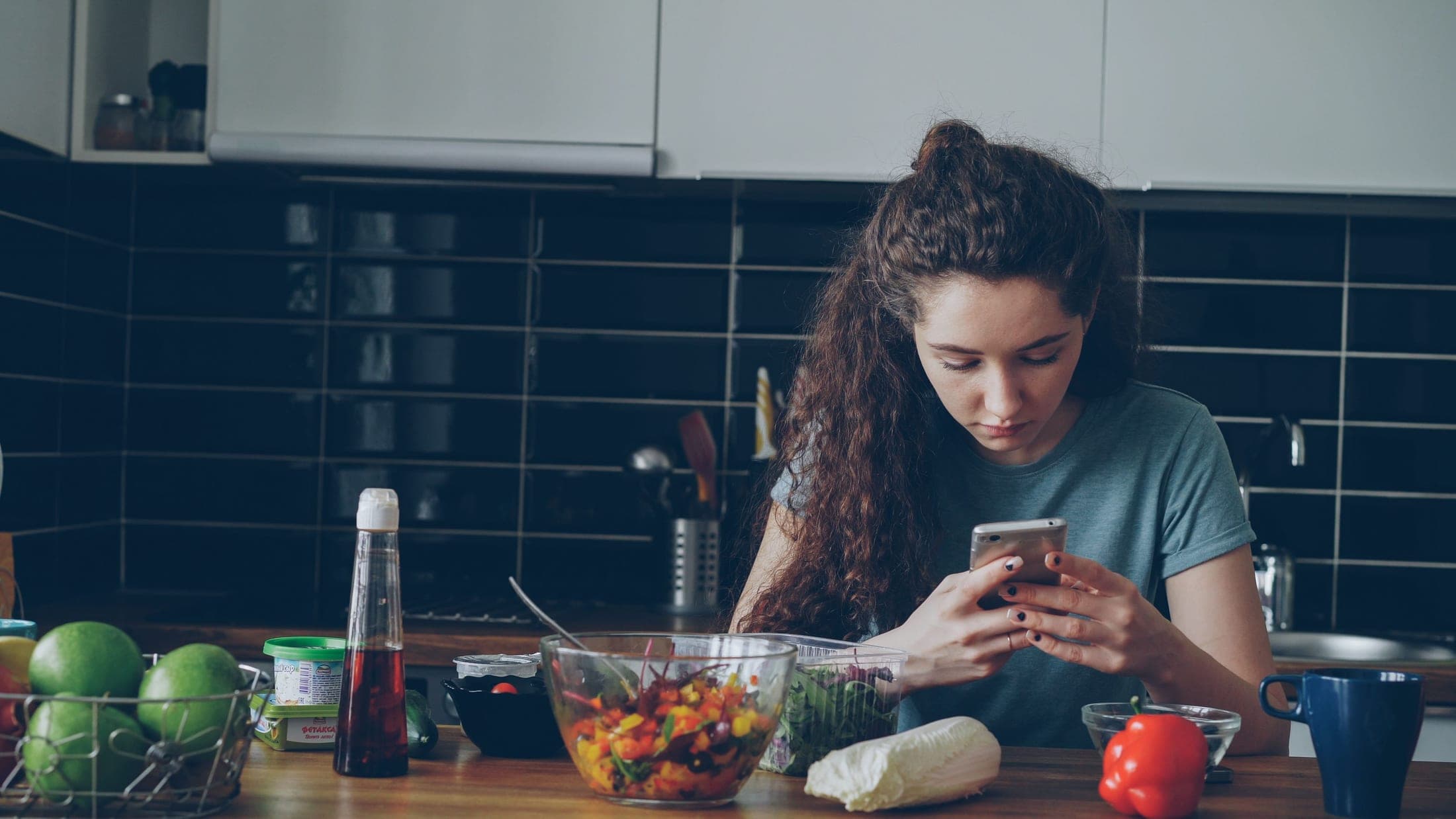 A woman at a kitchen counter surrounded by fresh produce and a salad she's prepared, looking at her phone.
