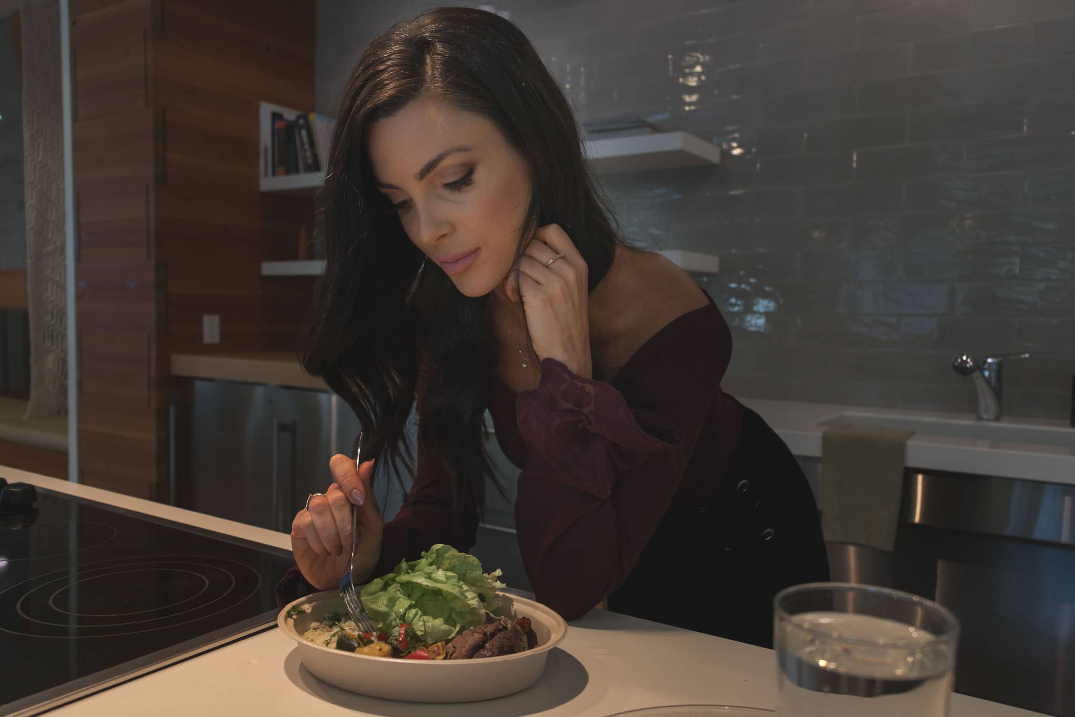 A woman at a kitchen island eating a fresh salad, fork in hand, focused on her food.